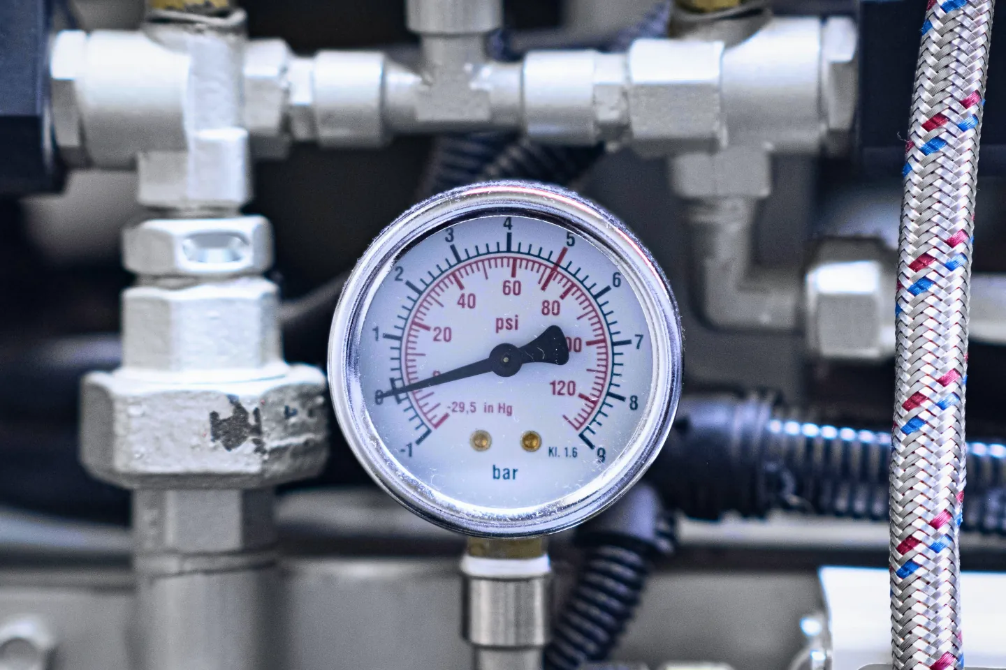 Manômetro com escala em bar e em psi, de −1 a 9 bar, instalado entre conexões roscadas de aço, com uma mangueira de malha trançada ao lado