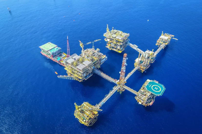 Complexo de plataformas de petróleo ligadas por passarelas, visto do alto, no mar azul, com um navio-guindaste atracado ao lado