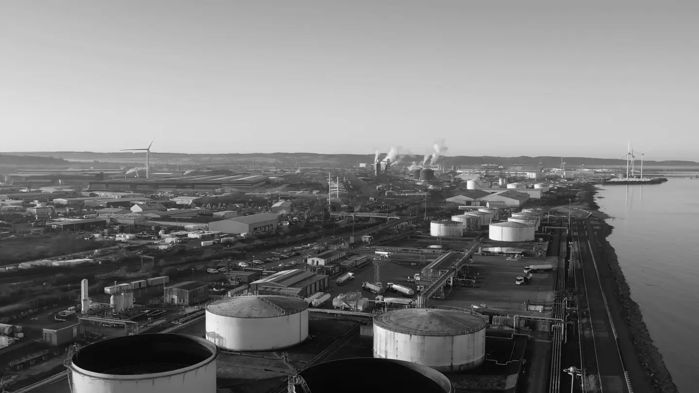 Vista aérea em preto e branco de um terminal de armazenamento à beira de um rio, com grandes tanques cilíndricos, caminhões-tanque e tubovias