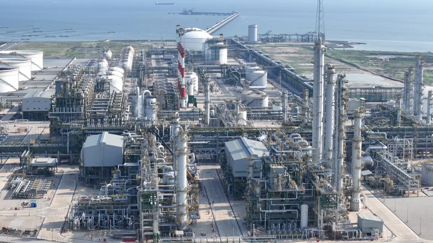Vista aérea das unidades de processo de uma refinaria à beira-mar, com colunas de destilação, racks de tubulação, tanques e uma chaminé listrada de vermelho e branco